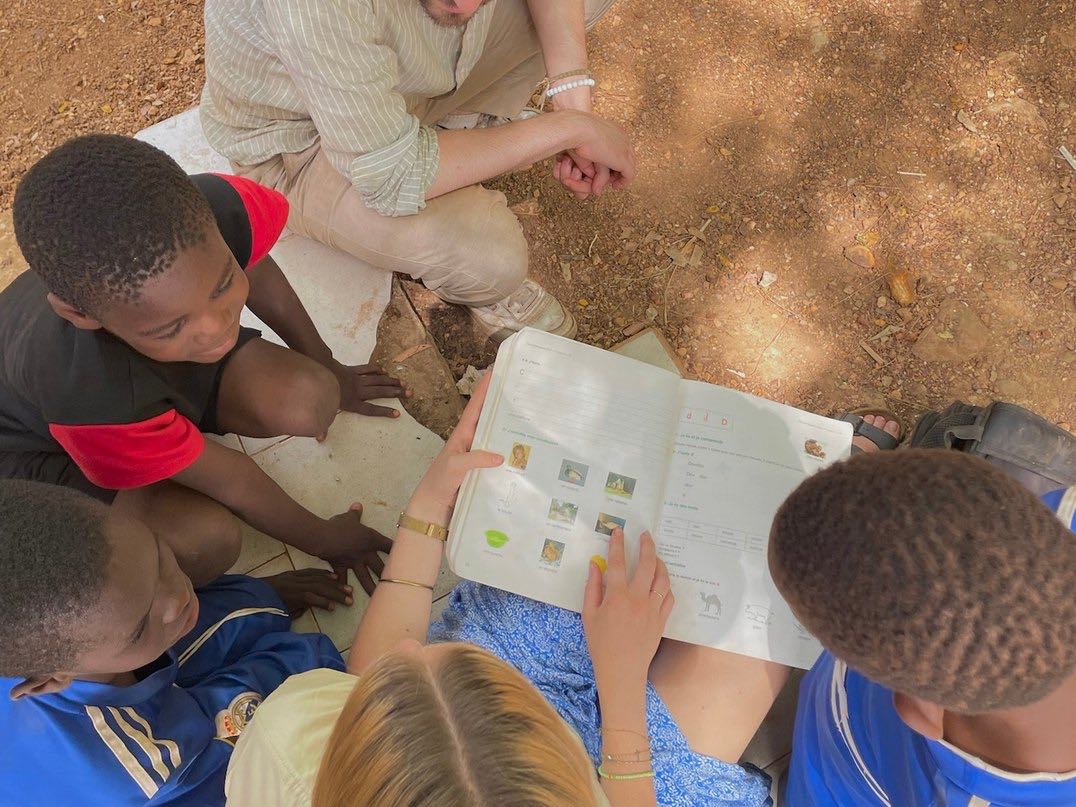 A volunteer reads a schoolbook with children sitting on the ground in Senegal